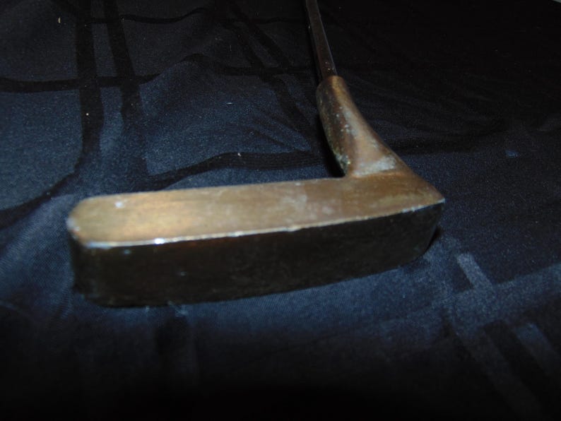 以下が含まれることがあります： ブロンズ色のヘッドと暗いシャフトが付いた、ヴィンテージのゴルフパター。パターヘッドは長方形です。クラブは使用感があり、摩耗や経年劣化の兆候が見られます。