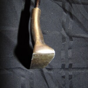 以下が含まれることがあります： ヴィンテージゴルフクラブのパターヘッドのクローズアップ。 パターヘッドは、くすんだ真鍮色の金属でできており、湾曲したネックと平らな長方形のフェースが付いています。 シャフトは濃い茶色です。