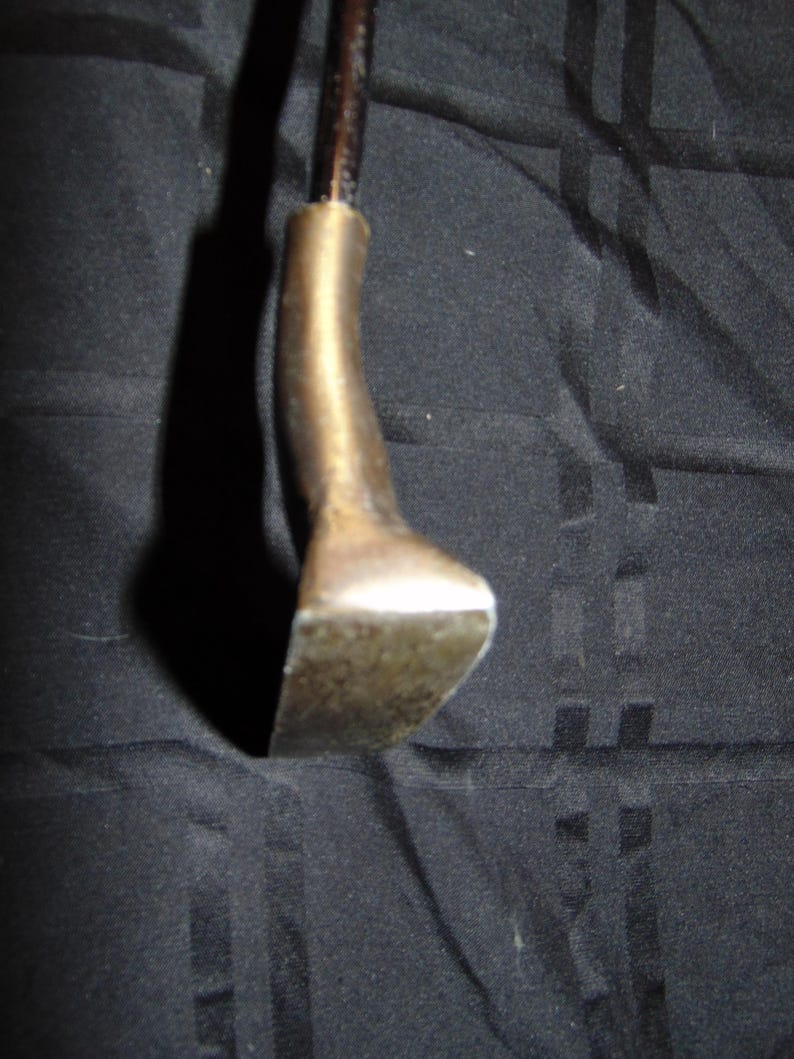 以下が含まれることがあります： ヴィンテージゴルフクラブのパターヘッドのクローズアップ。 パターヘッドは、くすんだ真鍮色の金属でできており、湾曲したネックと平らな長方形のフェースが付いています。 シャフトは濃い茶色です。