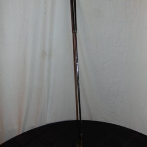 以下が含まれることがあります： 濃い茶色のシャフトと黒いグリップが付いたゴルフパター。パターヘッドは明るい茶色です。パターは黒い表面に置かれています。