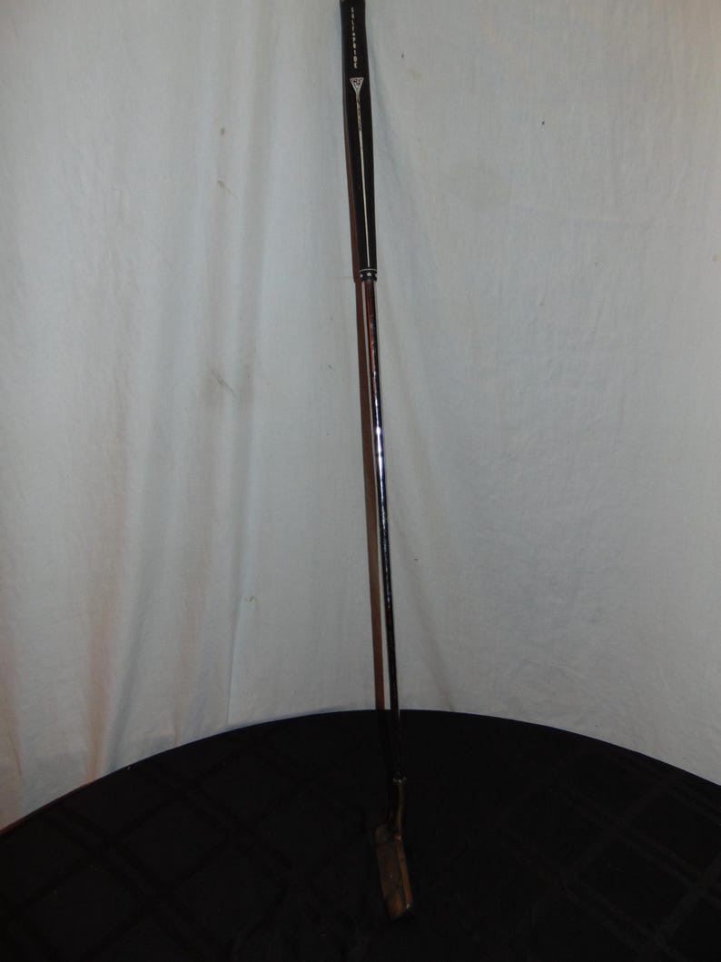 以下が含まれることがあります： 濃い茶色のシャフトと黒いグリップが付いたゴルフパター。パターヘッドは明るい茶色です。パターは黒い表面に置かれています。