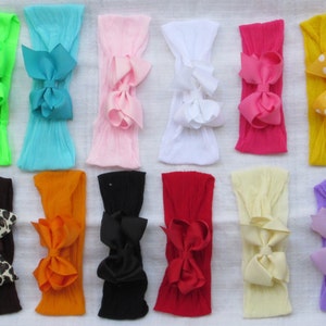 May include: A collection of colorful baby headbands. The headbands are in various colors, including green, blue, pink, white, red, yellow, brown, orange, black, and purple. Each headband features a large bow in a coordinating color. The headbands are arranged on a white surface.