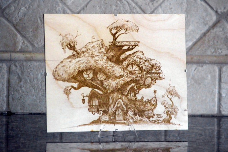 May include: A wood-burned image of a treehouse with multiple levels and windows. The treehouse is surrounded by trees and has a sign that reads "Fairy Circle".