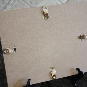 May include: The back of a brown picture frame with four metal hanging clips and a black easel stand.