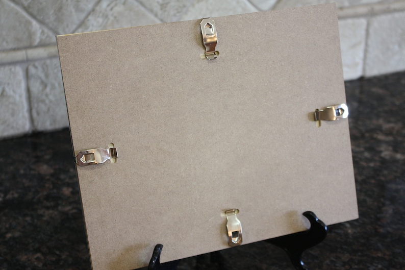 May include: The back of a brown picture frame with four metal hanging clips and a black easel stand.