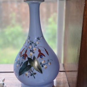 May include: A light blue vase with a bulbous base and a slender neck. The vase is decorated with hand-painted red, white, and green floral designs. The vase is displayed on a wooden shelf.