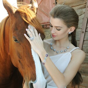 May include: A woman with long brown hair is gently touching a brown and white horse. She is wearing a white dress with a silver necklace and bracelet. The necklace has a floral design and the bracelet has round, multi-colored stones.