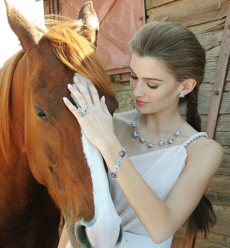 May include: A woman with long brown hair is gently touching a brown and white horse. She is wearing a white dress with a silver necklace and bracelet. The necklace has a floral design and the bracelet has round, multi-colored stones.