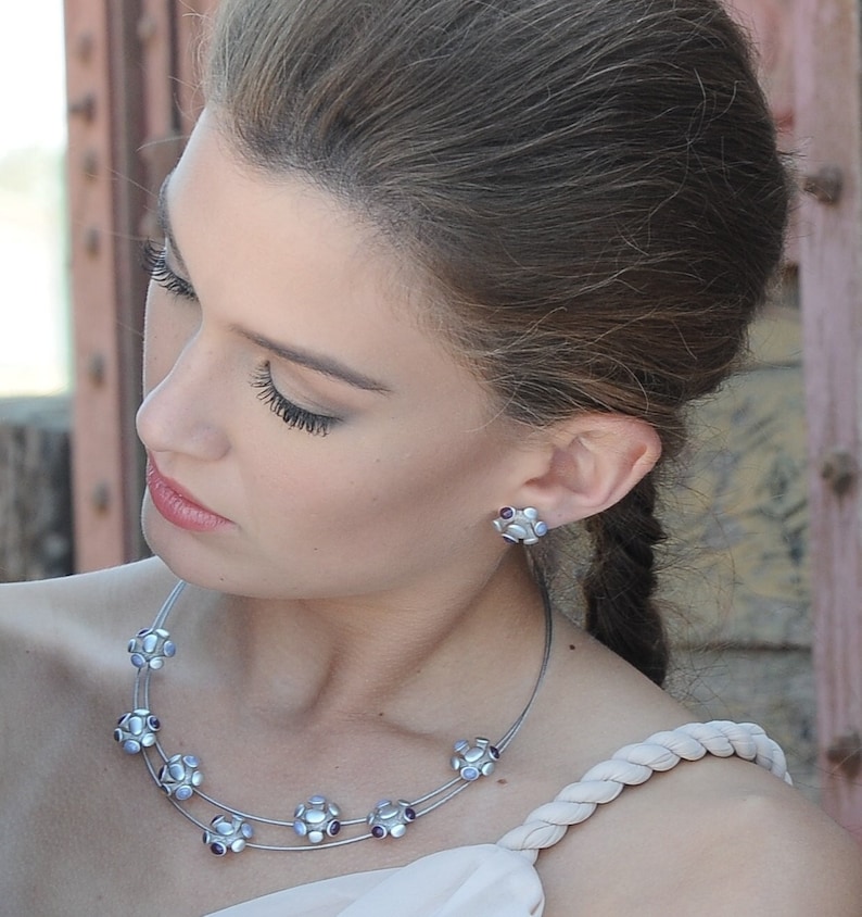 May include: A woman wearing a silver necklace with purple and white stones. The necklace has a delicate chain and is adorned with multiple flower-shaped pendants. The woman is also wearing a silver earring with purple and white stones.