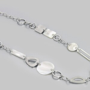May include: A silver chain necklace with white shell and metal geometric shapes. The necklace features a variety of shapes, including circles, squares, rectangles, and ovals.
