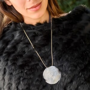 May include: A silver necklace with a large, round pendant featuring a spiral design. The pendant is hanging against a black fur background.