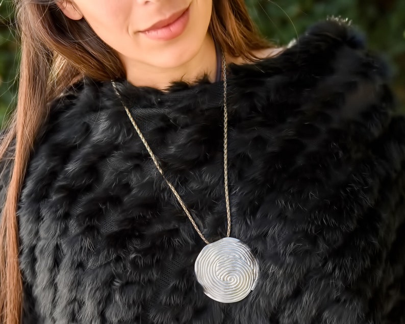 May include: A silver necklace with a large, round pendant featuring a spiral design. The pendant is hanging against a black fur background.
