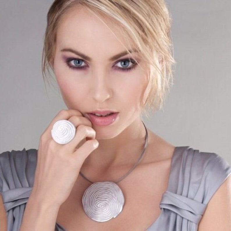 May include: A woman wearing a silver necklace and ring with a spiral design. The necklace and ring are both made of metal and have a smooth, polished finish.