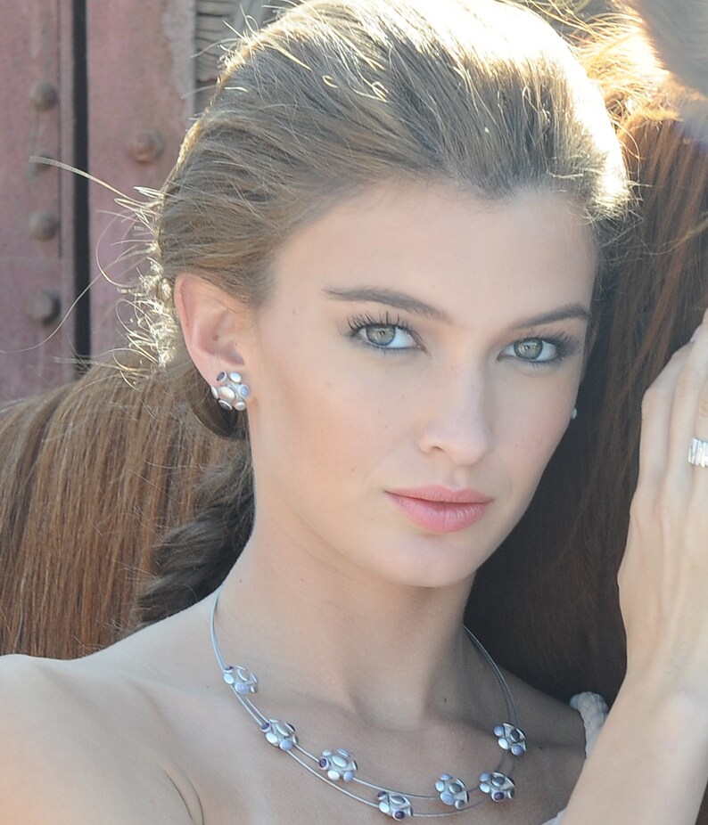 May include: A woman with long, light brown hair is wearing a silver necklace with purple stones. The necklace has a delicate, layered design.