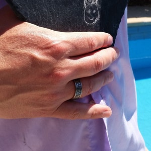 May include: A silver ring with a detailed pattern is worn on a hand. The ring has a dark, textured design. The hand is holding a dark, textured object, and is wearing a light purple shirt.