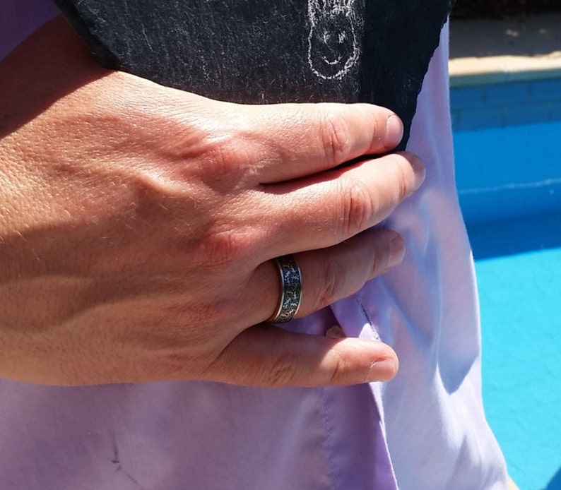 May include: A silver ring with a detailed pattern is worn on a hand. The ring has a dark, textured design. The hand is holding a dark, textured object, and is wearing a light purple shirt.