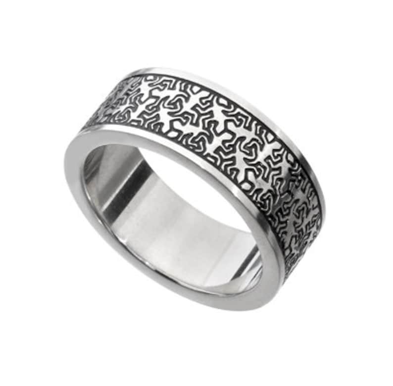 May include: A wide, silver-colored ring with a geometric pattern etched around the band. The pattern is a series of interconnected, abstract shapes in a darker tone, creating a contrast against the polished metal.