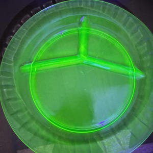 May include: A round, translucent, neon green glass plate featuring a three-section divided interior. The plate has a smooth central surface and a textured, ribbed rim. It glows with a bright, vibrant luminescence under ultraviolet light.