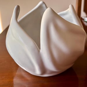 May include: A white ceramic bowl with a smooth, glossy finish and a sculptural design featuring four distinct, upward-curving petal-like sections that create a deep, open center. The object has a rounded base and a uniform, solid white color.