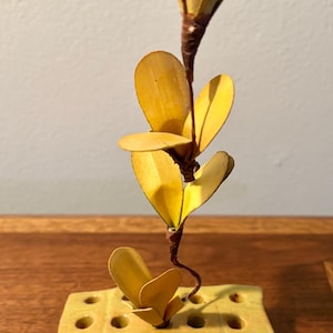 May include: A handcrafted, yellow ceramic flower frog with a decorative, artificial plant. The plant features yellow leaves and a brown stem. The flower frog has multiple holes for arranging stems. The piece is set on a wooden surface.