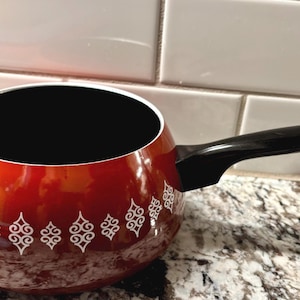 May include: A red fondue pot with a black interior and a black handle. The pot has a white decorative pattern around the exterior. The pot is sitting on a speckled countertop.