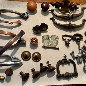 May include: A collection of various cabinet hardware, including handles, knobs, and pulls in different styles and finishes. The hardware includes silver, bronze, and wooden pieces, along with screws and other mounting hardware.