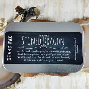 May include: A silver tin with a black label that reads "Unbound Stoned Dragon Beard Wax". The label has a humorous description of a dragon who got too baked and the text "www.etsy.com/shop/Unbound Elements".