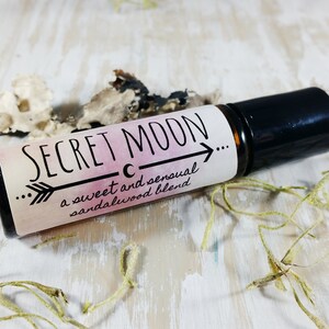 May include: A black roller bottle with a white label that says "SECRET MOON... a sweet and sensual sandalwood blend". The label has a crescent moon and an arrow pointing to the right.
