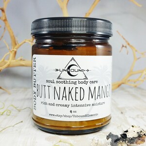 May include: A glass jar with a black lid containing body butter. The label reads "Unbound" and "Butt Naked Mango" with the tagline "Soul Soothing Body Care" and "Rich and Creamy Intensive Moisture". The jar is 4 oz and is available at www.etsy.com/shop/UnboundElements.