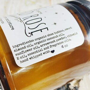 May include: An amber-coloured jar of body butter with a white label. The label reads "ROSE" and lists ingredients including shea butter and almond oil. The jar contains 113 grams of product.