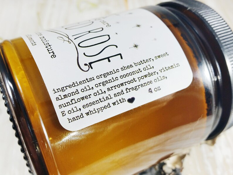 May include: An amber-coloured jar of body butter with a white label. The label reads "ROSE" and lists ingredients including shea butter and almond oil. The jar contains 113 grams of product.