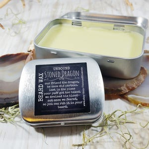 May include: A silver tin with a black label that reads "Beard Wax Unbound Stoned Dragon" and a description of the product. The tin is open and contains a yellow beard wax.