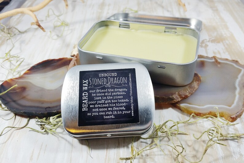 May include: A silver tin with a black label that reads "Beard Wax Unbound Stoned Dragon" and a description of the product. The tin is open and contains a yellow beard wax.