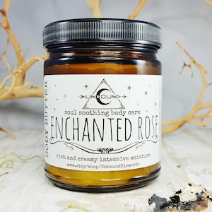 May include: An amber glass jar of body butter with a black lid. The label reads "ENCHANTED ROSE" with the text "soul soothing body care" and "rich and creamy intensive moisture". The jar is on a light-coloured surface.