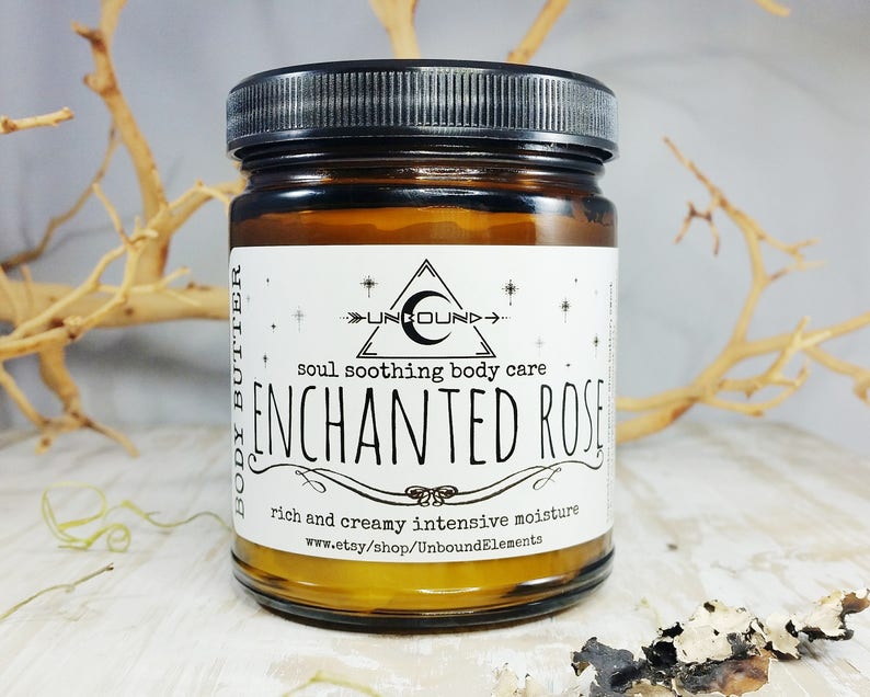 May include: An amber glass jar of body butter with a black lid. The label reads "ENCHANTED ROSE" with the text "soul soothing body care" and "rich and creamy intensive moisture". The jar is on a light-coloured surface.