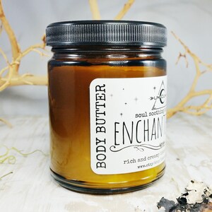 May include: A jar of body butter with a black lid and a white label. The label reads "BODY BUTTER" and "ENCHANT". The jar is filled with a golden-coloured cream. The background includes dried branches.
