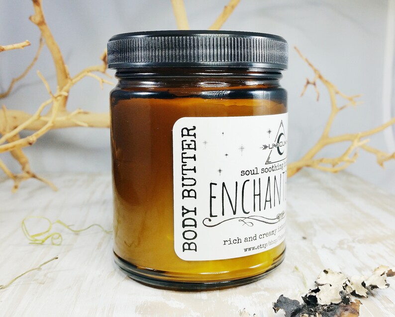May include: A jar of body butter with a black lid and a white label. The label reads "BODY BUTTER" and "ENCHANT". The jar is filled with a golden-coloured cream. The background includes dried branches.