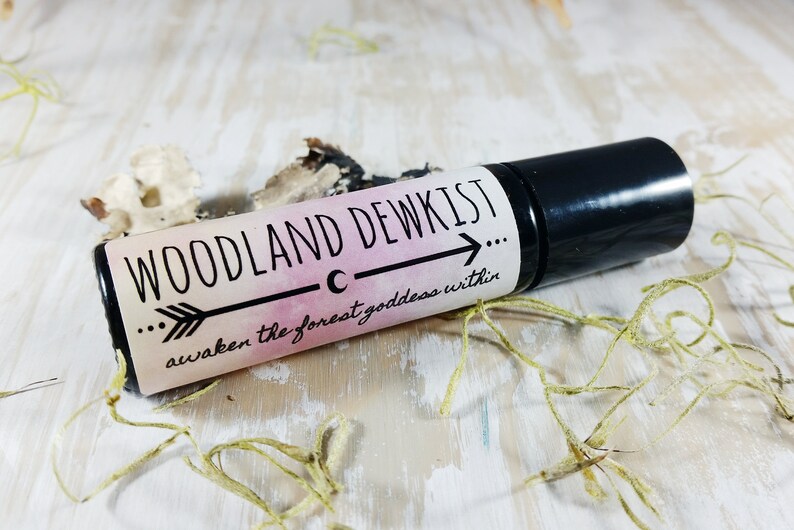 May include: A black roller bottle with a white label that reads "WOODLAND DEWKIST... awaken the forest goddess within..." The label has a pink and white watercolor background with a black arrow pointing to the right.