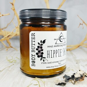 May include: A brown glass jar with a black lid containing body butter. The label reads "BODY BUTTER", "UNBOUND", "soul soothing body butter", "HIPPIE SKIN", "rich and creamy intensive", "4 oz", and "www.etsy.shop/unboundmoon".