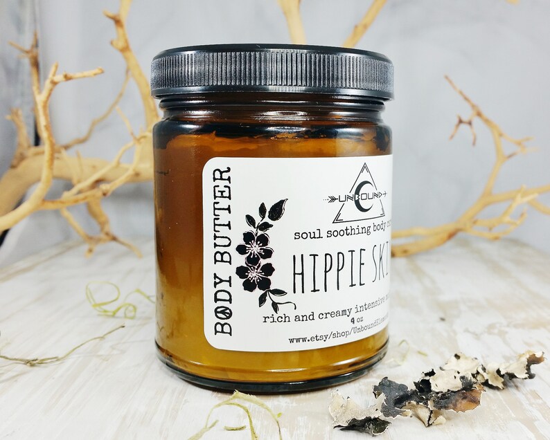 May include: A brown glass jar with a black lid containing body butter. The label reads "BODY BUTTER", "UNBOUND", "soul soothing body butter", "HIPPIE SKIN", "rich and creamy intensive", "4 oz", and "www.etsy.shop/unboundmoon".