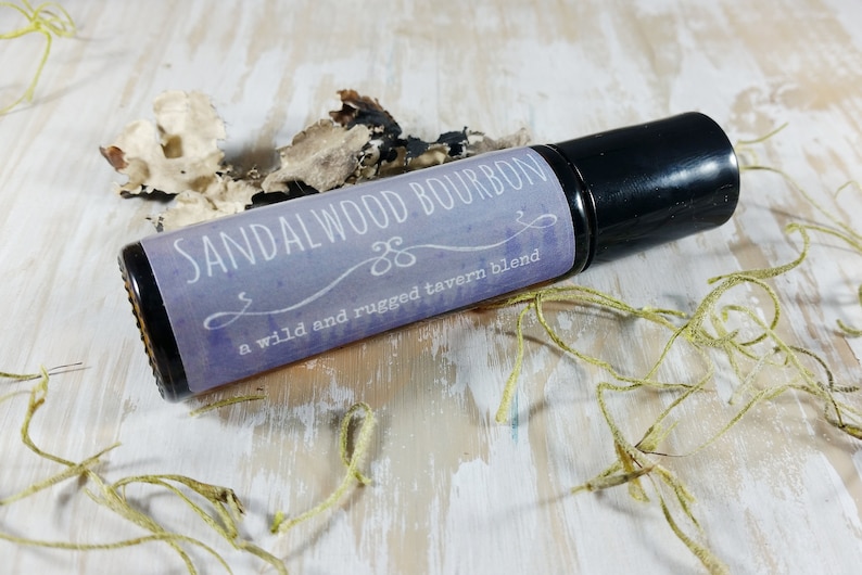 May include: A brown glass roller bottle with a black cap. The label is purple and white with the text "SANDALWOOD BOURBON" and "a wild and rugged tavern blend".
