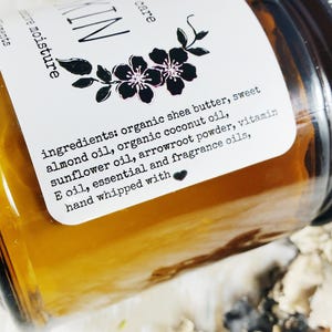 May include: A close-up of a glass jar with a brown label. The label reads "ingredients: organic shea butter, sweet almond oil, organic coconut oil, sunflower oil, arrowroot powder, vitamin E oil, essential and fragrance oils, hand whipped with".