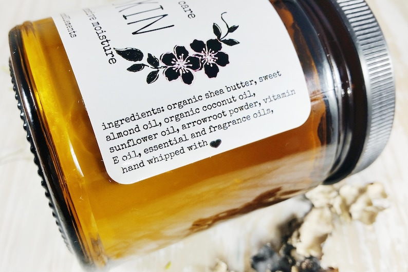 May include: A close-up of a glass jar with a brown label. The label reads "ingredients: organic shea butter, sweet almond oil, organic coconut oil, sunflower oil, arrowroot powder, vitamin E oil, essential and fragrance oils, hand whipped with".