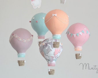 May include: A baby mobile with five hot air balloons in pink, peach, light blue, and gray with white accents. The balloons are suspended from a white metal rod with a white cloud and a string of flags in pink, peach, and green.
