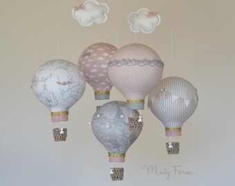 May include: A mobile with five hot air balloons in pink, gray, and white fabric with different patterns. The balloons are suspended from a white ceiling with white clouds. The balloons have gold trim and are attached to small baskets.
