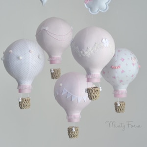 Op de afbeelding: Een roze en witte babymobiel met vijf luchtballonnen. Elke ballon heeft een ander ontwerp, waaronder stippen, kant en bloemenpatronen. De ballonnen hangen aan een witte wolk.
