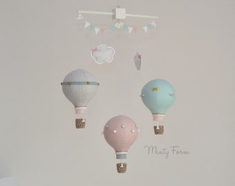 May include: A baby mobile featuring three hot air balloons in pastel colors. The balloons are striped, polka-dotted, and dotted, with woven baskets. A banner and cloud accents complete the nursery decor.