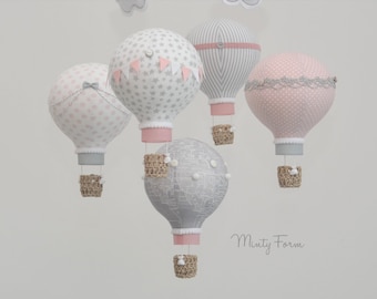 May include: A mobile with five hot air balloons in pink and gray with white polka dots, stars, and stripes. The balloons are suspended from a white ceiling with a gray background. The balloons have wicker baskets.