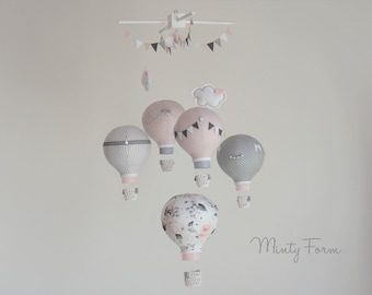 May include: A baby mobile with five hot air balloons in pink, gray, and white fabric patterns. The balloons are suspended from a white wooden frame with a pink and white fabric banner.
