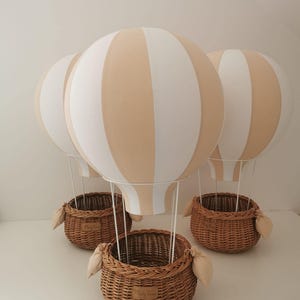 May include: Three decorative hot air balloon ornaments featuring round, beige and white striped fabric balloons attached to woven brown wicker baskets by thin white wires. Each basket includes small fabric side accents and a small label.